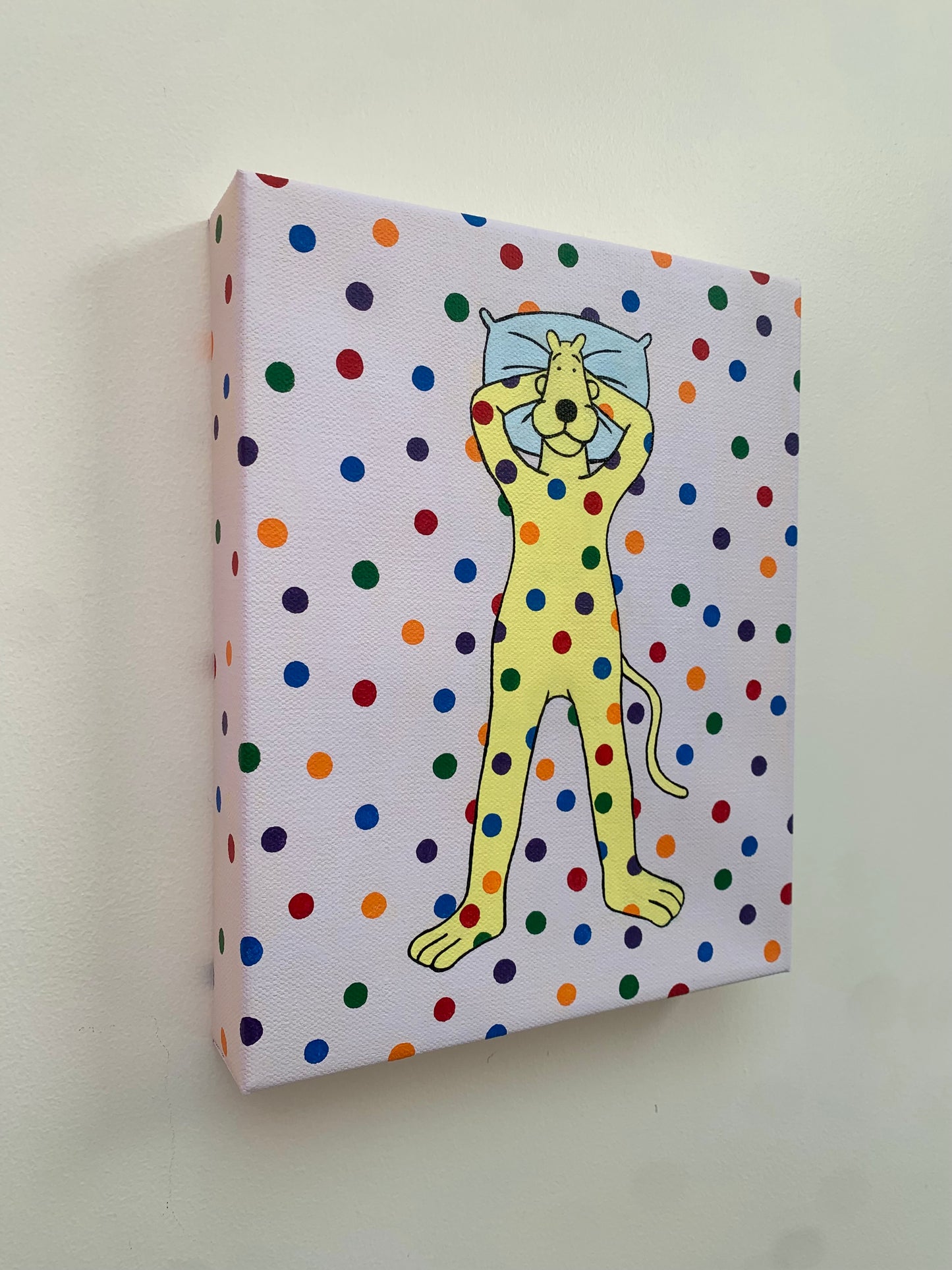 Canvas painting of a yellow cartoon leopard lying on the ground with his head on a pillow. Rainbow spots are all over him and the background