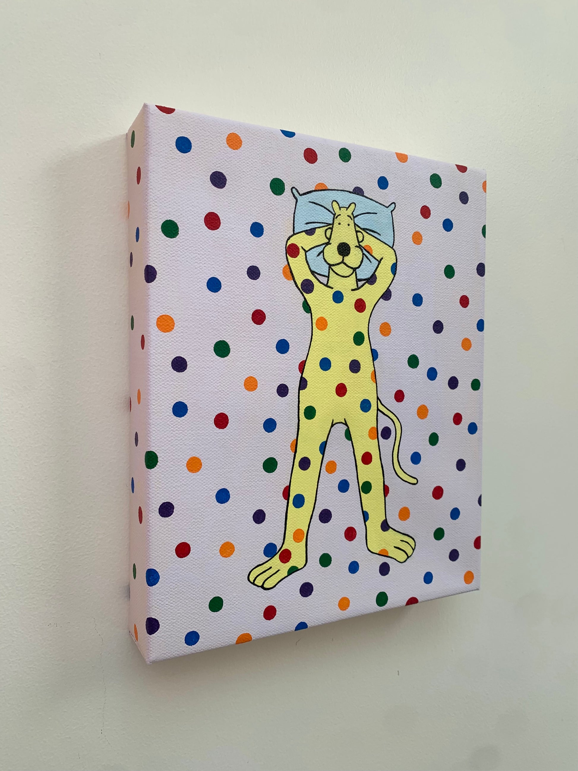 Canvas painting of a yellow cartoon leopard lying on the ground with his head on a pillow. Rainbow spots are all over him and the background