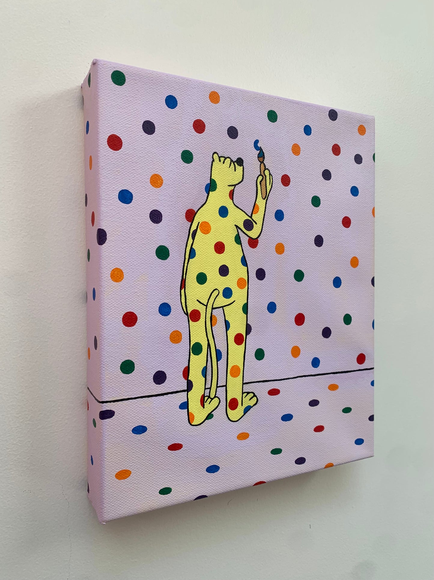 Canvas painting on a white wall of the yellow leopard covered in rainbow spots standings with his back facing us. He has a paintbrush in hand and is painting the spots on the wall.