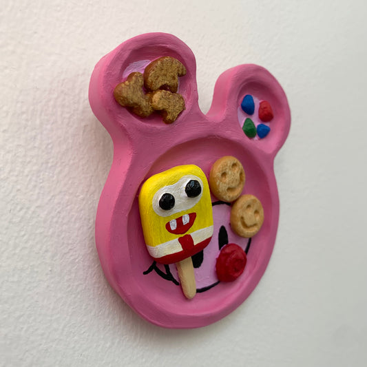 Pink mouse shaped sculpture on a white wall with rainbow dessert treats on the surface