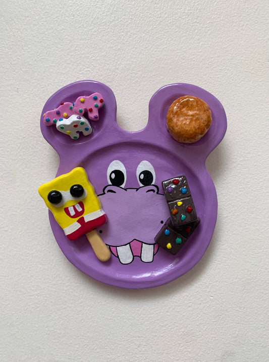 Purple hippo-shaped plate with cartoon face and various items on a white background